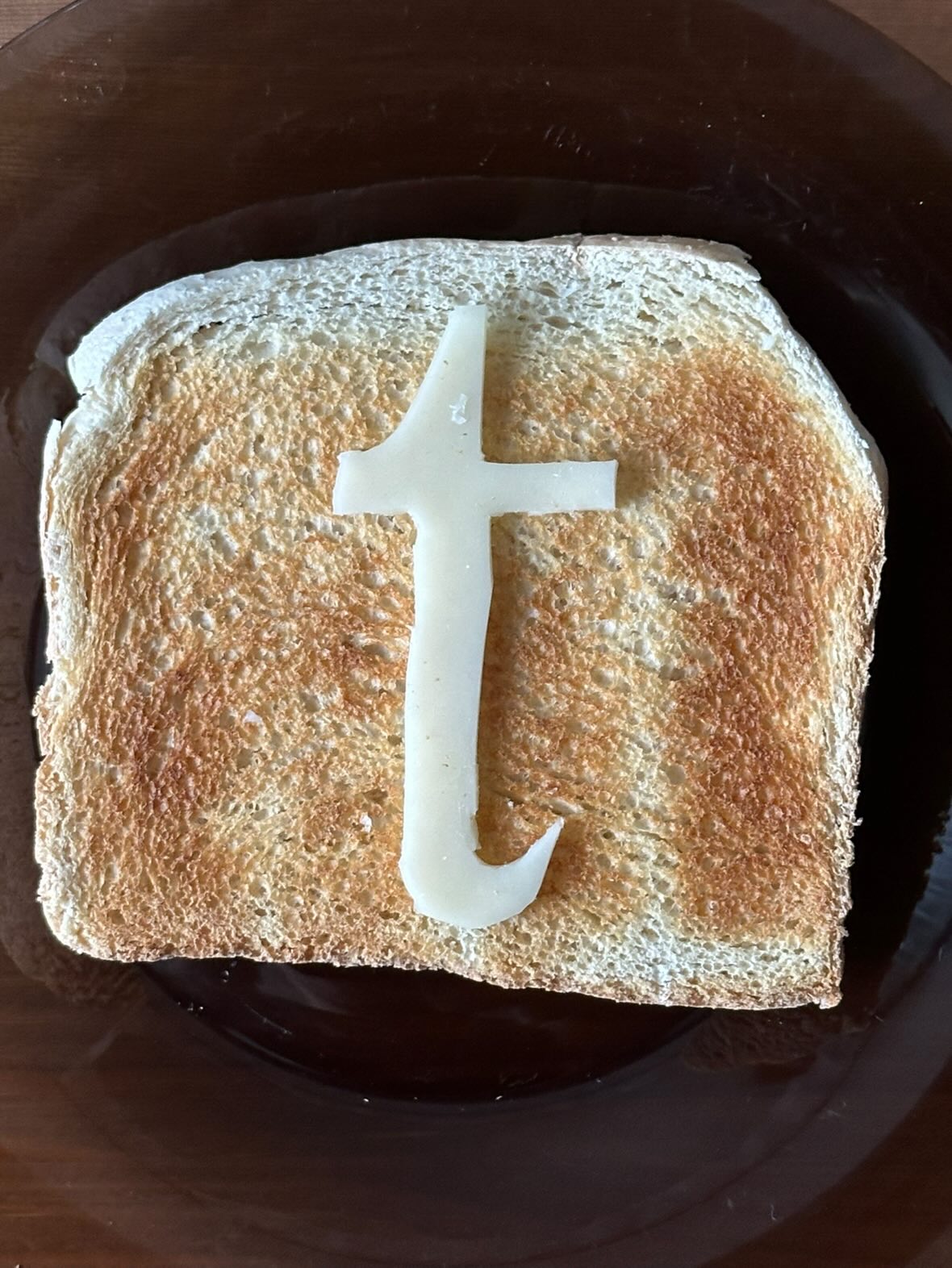 A slice of toasted bread on a dark plate with a cheese cutout shaped like a lowercase “t” placed neatly on top. 